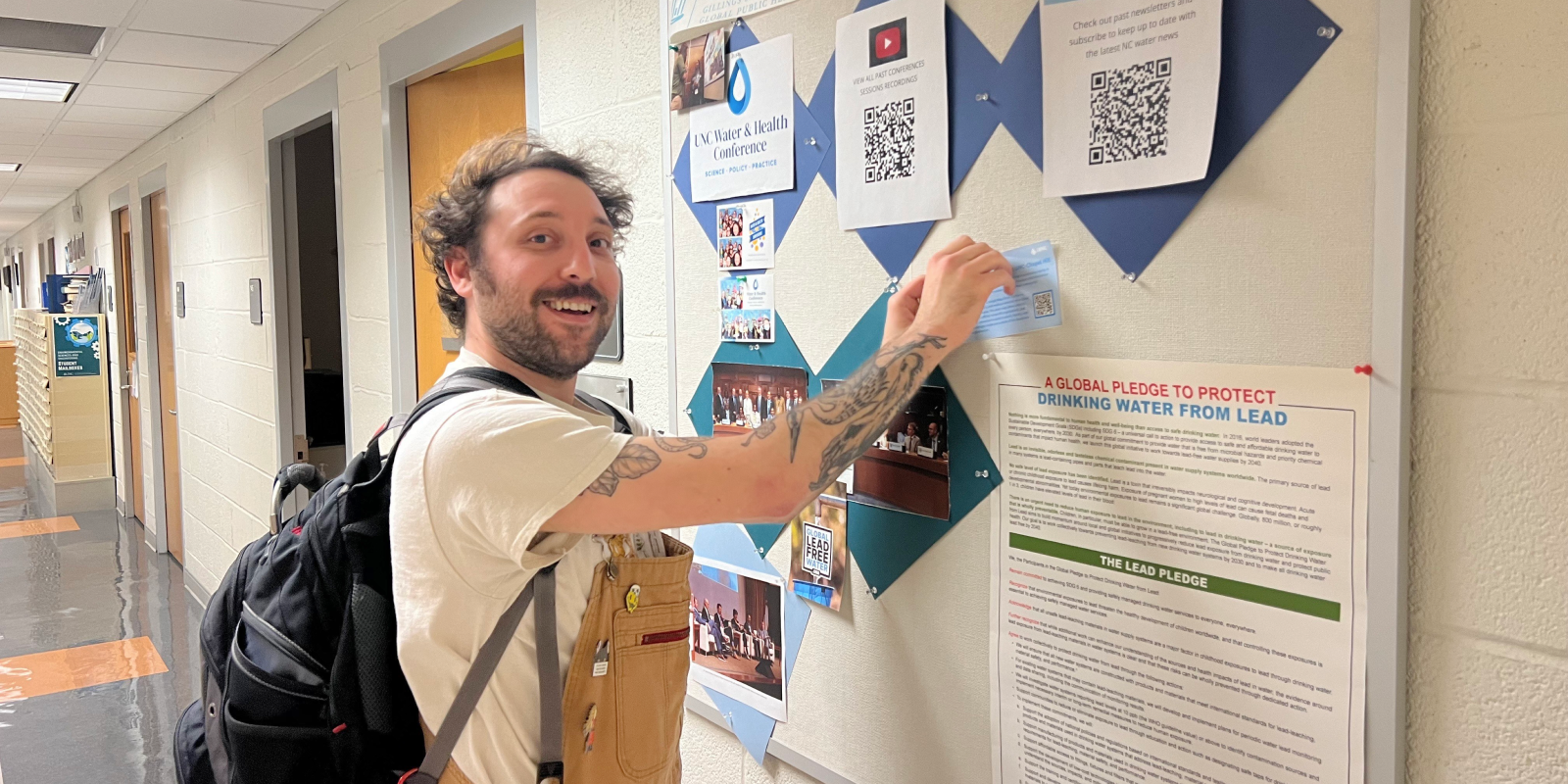 A student with a beard and tattoos on his arm is standing in a hallway, smiling at the camera. He is wearing a white t-shirt and brown overalls with a black backpack. He is pinning an mRelief flyer to a bulletin board covered with various posters and flyers, including one about protecting drinking water from lead and a QR code for a conference. The hallway has a light-colored tiled floor and several doors along the walls.