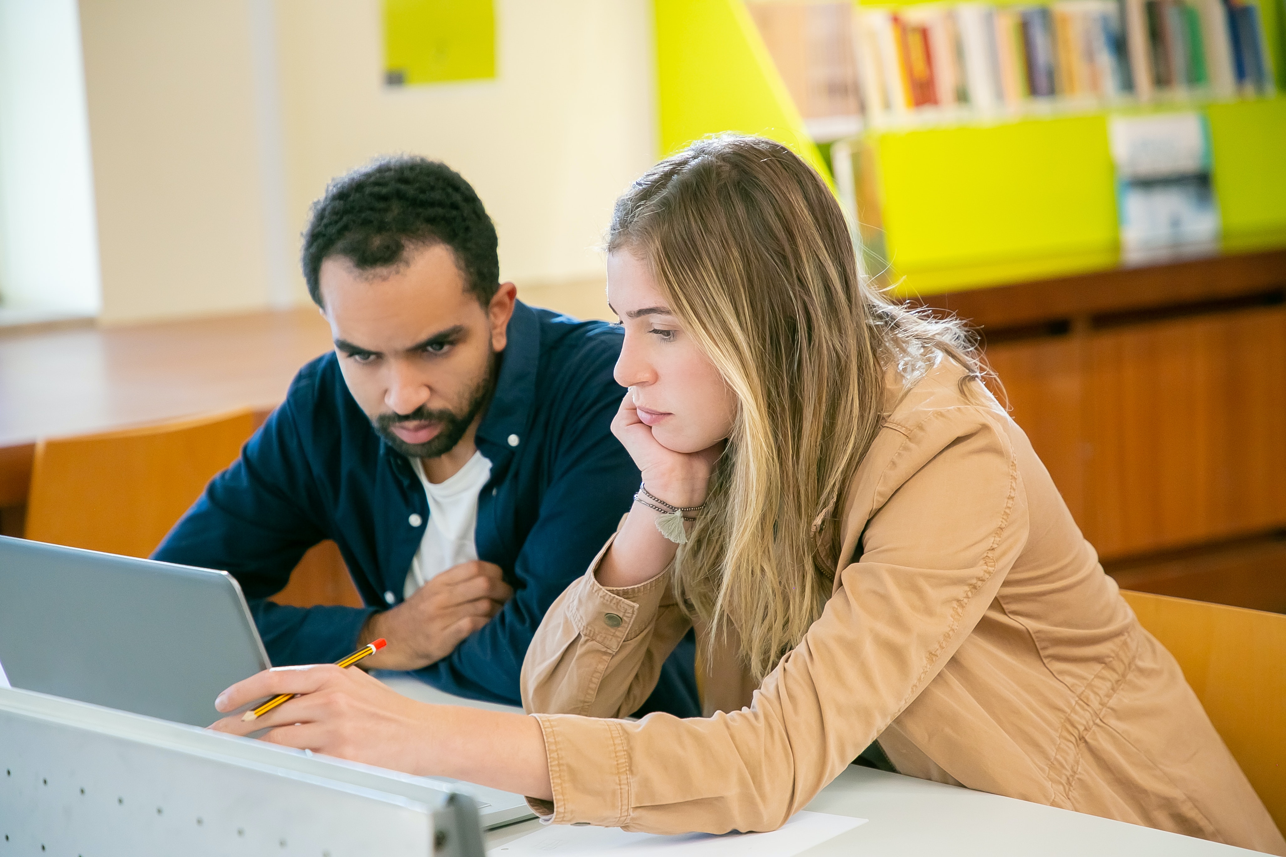 A young adult man with a short dark beard and short dark hair and olive skin wearing a long blue button down shirt unbuttoned over a white tshirt is seated next to a young adult woman with long blonde hair and fair skin wearing a khaki jacket. They are looking at a computer together while seated at a grey table in a well-lit, modern library with beige walls and bright yellow bookshelves.