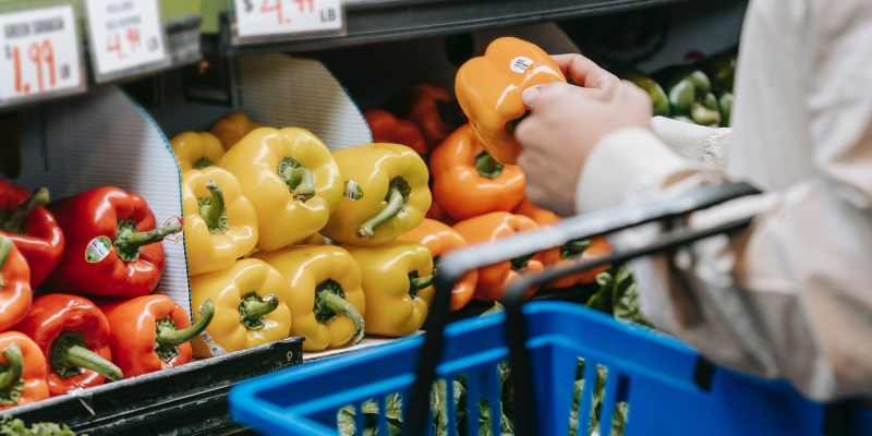 A photo of a row of bell peppers at a supermarket and someone putting one orange bell pepper into a shopping basket.