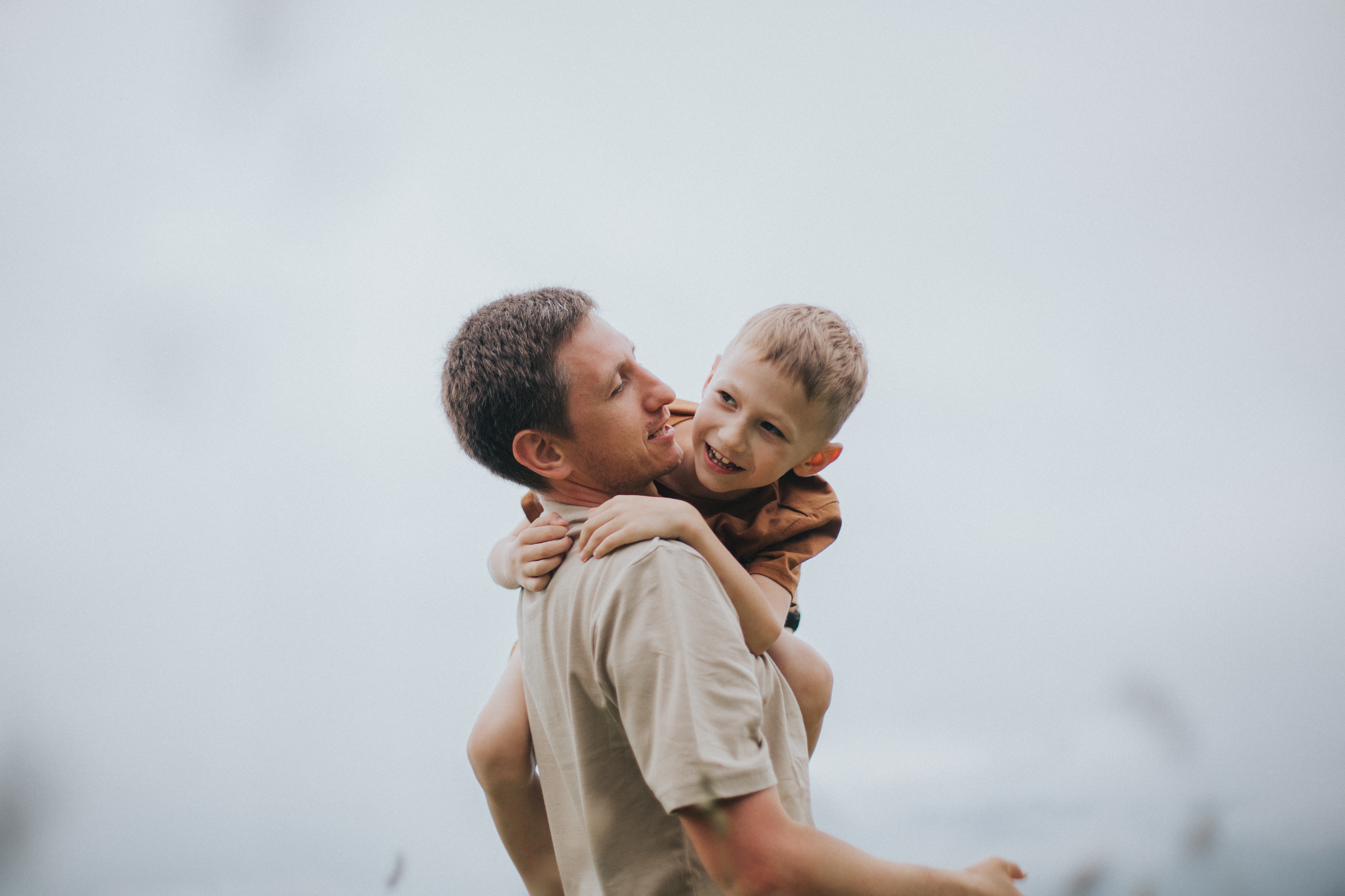 A photo of a young child and man smiling while the child is being carried on a man's shoulders with their arms wrapped around his back.